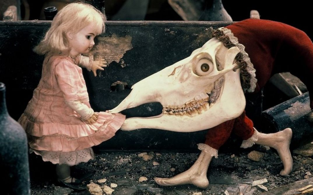 Filmstill aus ALICE: Eine Puppe mit blondem Haar und rosafarbenen Kleid, neben ihr eine groteske Figur, mit einem Tierschädel, der das Kleid in den Mund nimmt.