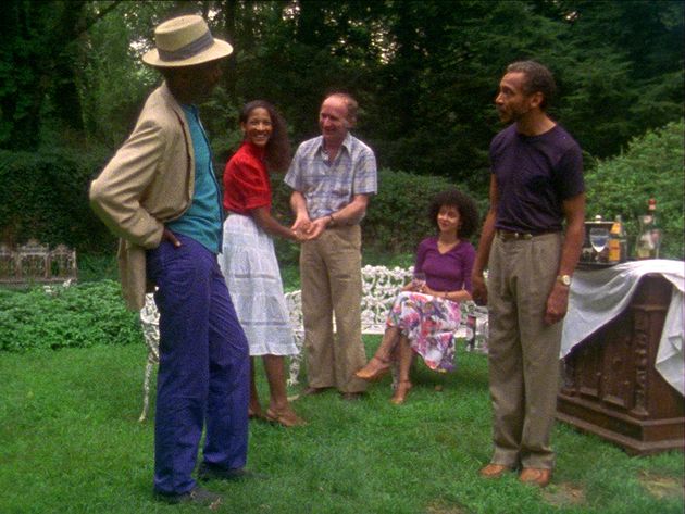 Still from the film „Losing Ground“. A group of people of different ages is standing on the grass in a garden, looking at each other and seemingly in a good mood. 