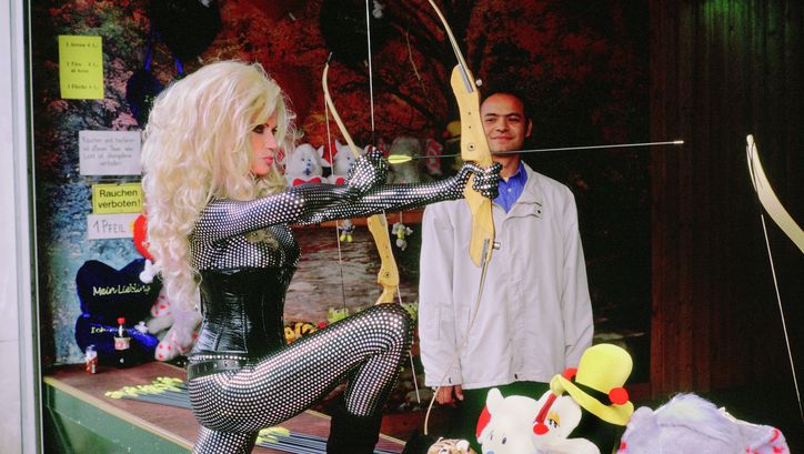 Film still from PRATER: A blonde woman wearing a tight bodysuit and corsage is standing shooting gallery holding a bow and arrow.