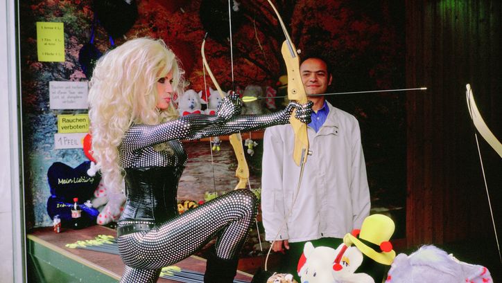 Film still from PRATER: A blonde woman wearing a tight bodysuit and corsage is standing shooting gallery holding a bow and arrow.