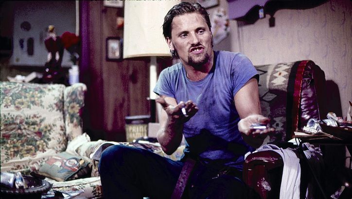 Filmstill aus SALVATION!: Ein Mann in blauem Shirt und Jeans sitzt auf einem Sessel und gestikuliert stark mit seinen Händen. Er spricht aufgebraust. Im Hintergrund ein Wohnzimmer.