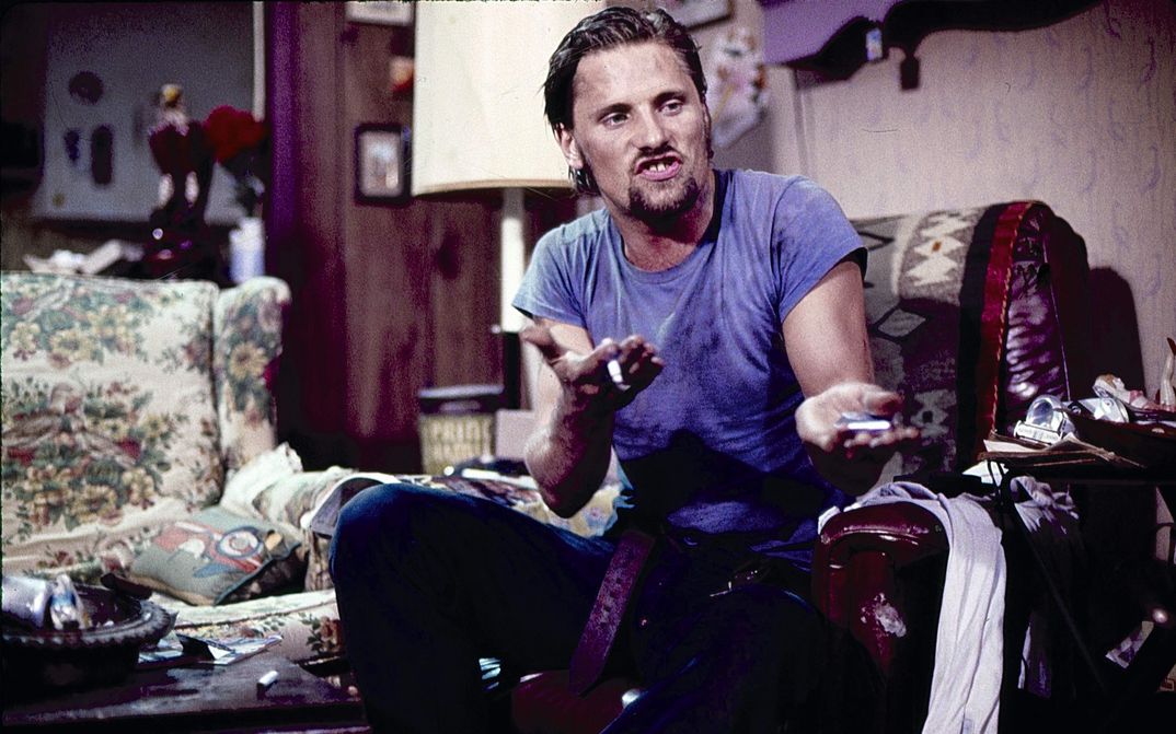 Filmstill aus SALVATION!: Ein Mann in blauem Shirt und Jeans sitzt auf einem Sessel und gestikuliert stark mit seinen Händen. Er spricht aufgebraust. Im Hintergrund ein Wohnzimmer.