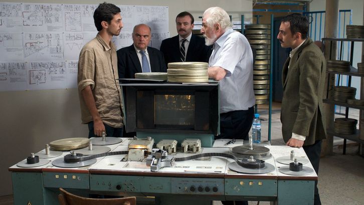 Film still from CHANTRAPAS: A group of men discussing in a room. An editing table with a 35mm film can be seen in the foreground. The room is full of film cans and a drawn storyboard is hanging.