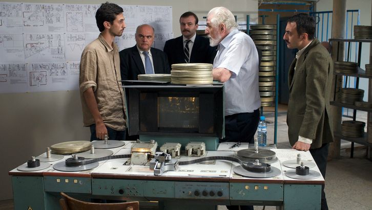 Film still from CHANTRAPAS: A group of men discussing in a room. An editing table with a 35mm film can be seen in the foreground. The room is full of film cans and a drawn storyboard is hanging.