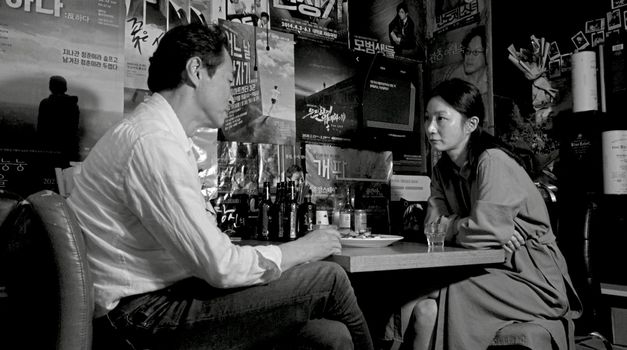 Black and white image of a man and a woman sitting opposite each other at a table with food and drink. The wall behind the table is covered with posters.
