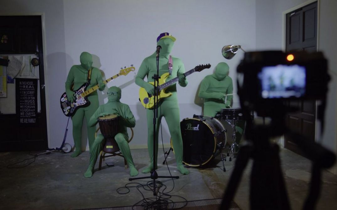 A rock band in green full-body suits, which also cover their heads, stand in formation in front of a digital camera. They hold their instruments in their hands.
