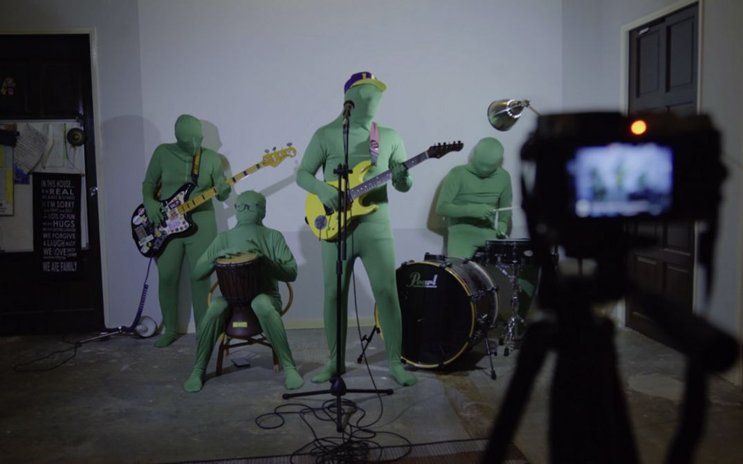 A rock band in green full-body suits, which also cover their heads, stand in formation in front of a digital camera. They hold their instruments in their hands.