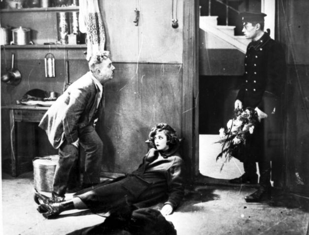 Film still from DAS MÄDCHEN MIT DER HUTSCHACHTEL: A young woman is half lying and half sitting, leaning against the wall, in an apartment. To her left is an older man looking at a younger man in uniform standing in the doorway with a bouquet of flowers in his hand.