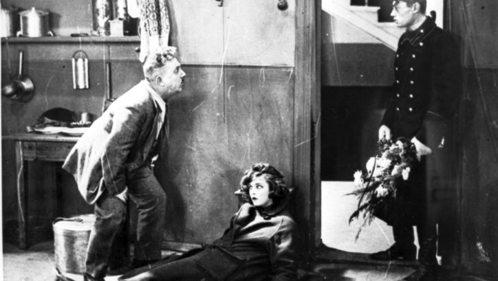 Film still from DAS MÄDCHEN MIT DER HUTSCHACHTEL: A young woman is half lying and half sitting, leaning against the wall, in an apartment. To her left is an older man looking at a younger man in uniform standing in the doorway with a bouquet of flowers in his hand.