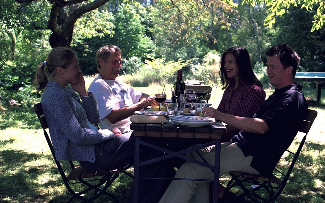 Two men and two women sit at a garden table set with wine and food and look happy.