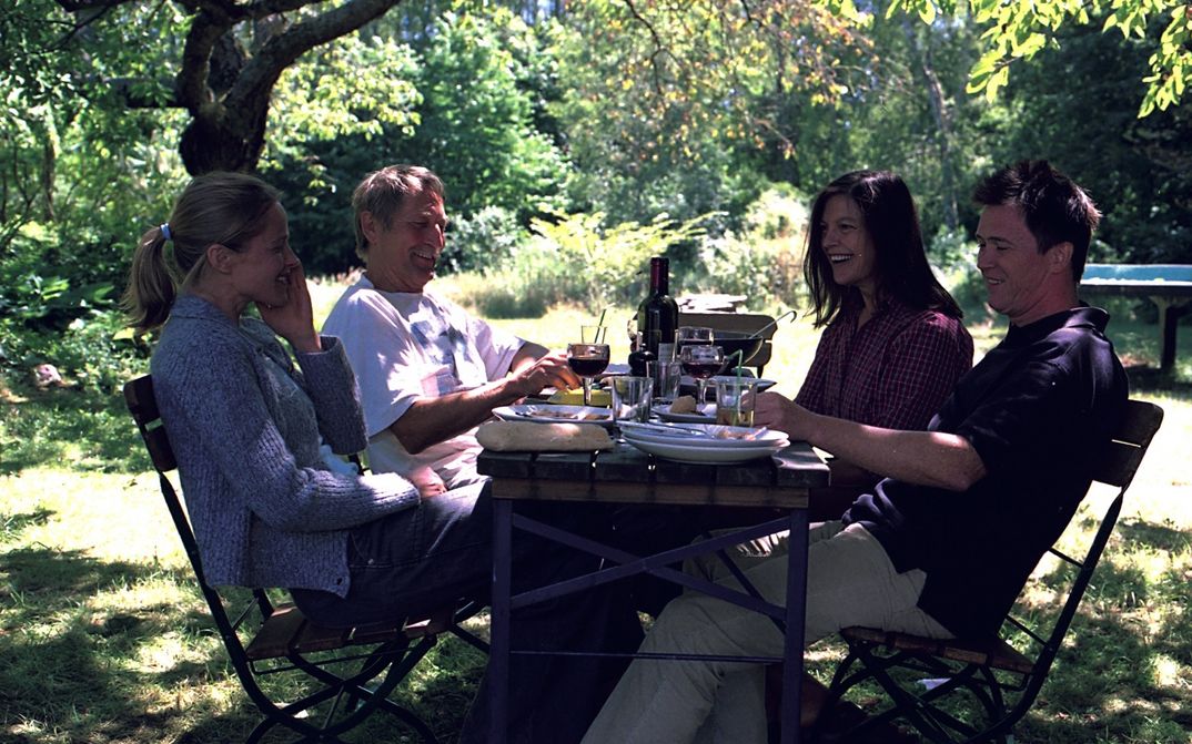 Two men and two women sit at a garden table set with wine and food and look happy.