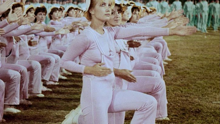Still from the film "Între revoluții" by Vlad Petri. Two rows of women in pink outfits are kneeling next to each other, their left arms extended forwards. There is a blonde woman with a light hat in the centre of the image. 