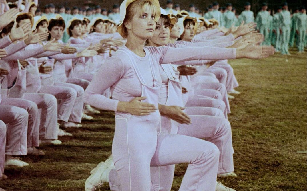 Still from the film "Între revoluții" by Vlad Petri. Two rows of women in pink outfits are kneeling next to each other, their left arms extended forwards. There is a blonde woman with a light hat in the centre of the image. 
