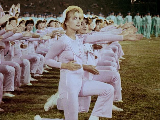 Still from the film "Între revoluții" by Vlad Petri. Two rows of women in pink outfits are kneeling next to each other, their left arms extended forwards. There is a blonde woman with a light hat in the centre of the image. 