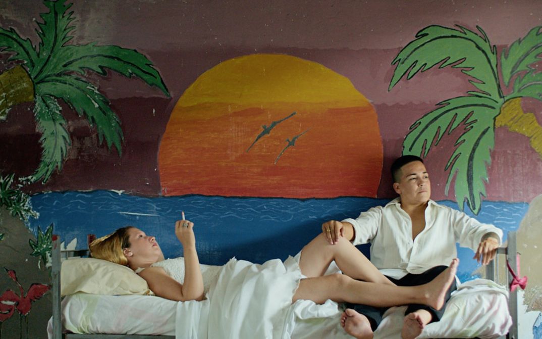 Filmstill aus "Reas" von Lola Arias. Zu sehen ist eine rauchende Frau, die auf einem Bett liegt, und ein Mann, der am Fußende des Bettes sitzt. Die Wand ist mit einem Sonnenuntergang bemalt.