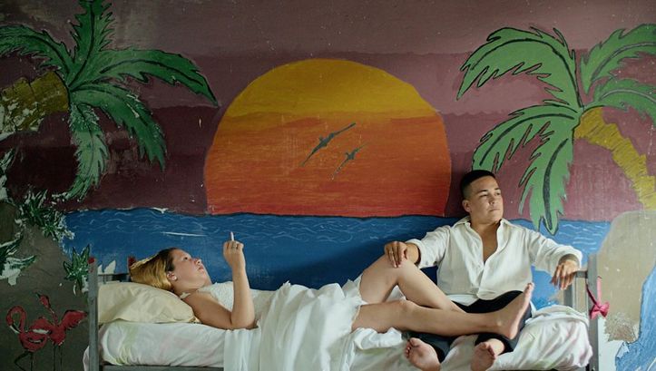 Film still from "Reas" by Lola Arias. It shows a smoking woman lying on a bed and a man sitting at the foot of the bed. The wall is painted with a sunset.