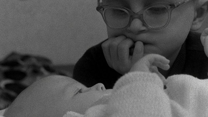 Film still from WHERE DO BABIES COME FROM: A small child with glasses looks at a baby lying in front of him.