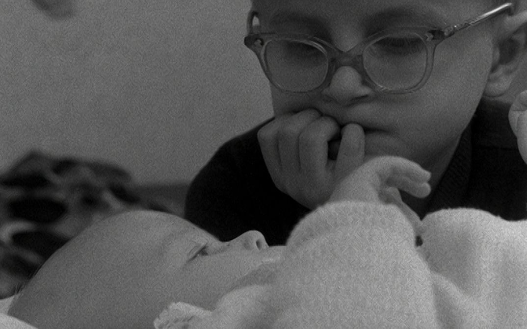 Filmstill aus WHERE DO BABIES COME FROM: Ein kleines Kind mit Brille betrachtet ein Baby, das vor ihm liegt.
