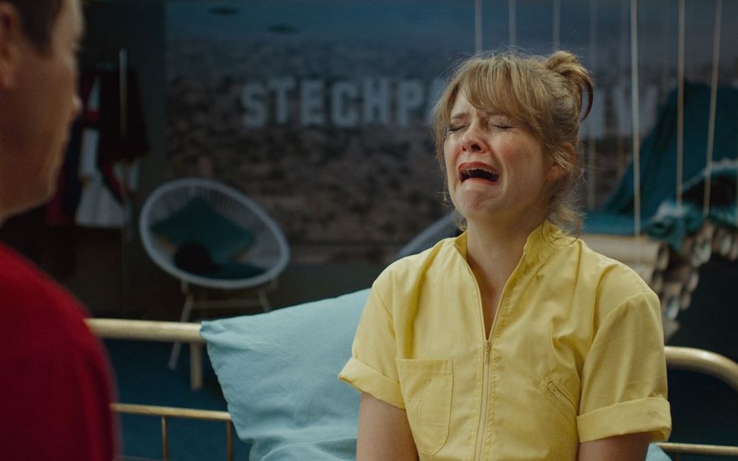 Film still from ZWEI IM FALSCHEN FILM: Laura Tonke, wearing a yellow top, is crying in a theatrical manner.