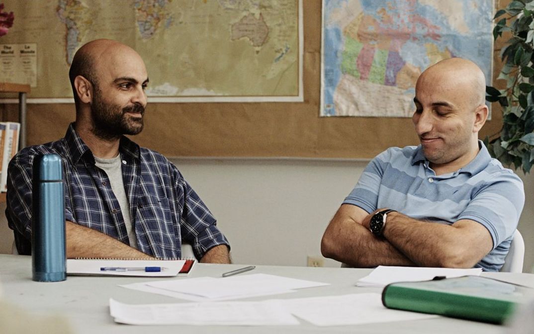 Filmstill from „Concrete Valley" by Antoine Bourges. Two men are sitting at a table in a classroom and are talking happily.