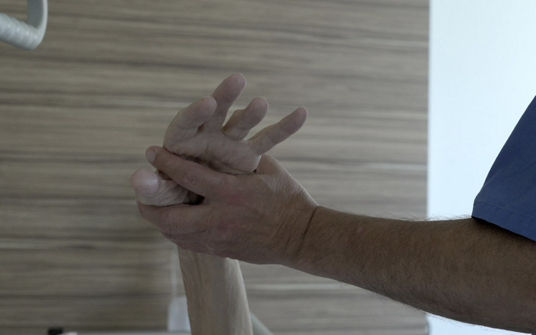 Ein Arm einer Krankenpfleger*in hält die Hand eine*r Patient*in hoch vor einer Wand mit Holzfournier.