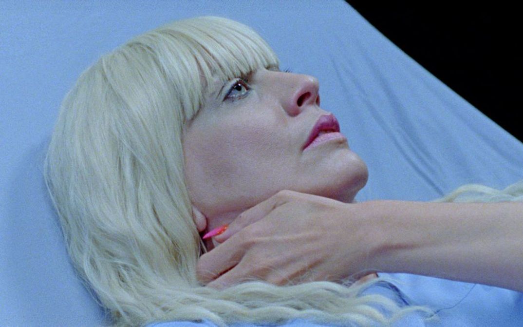 Filmstill aus SIRENS CALL von Miri Ian Gossing und Lina Sieckmann. Das Bild zeigt eine Nahaufnahme einer blonden Frau in einem Krankenbett. Mit ihrer Hand fasst sie sich an den Hals.