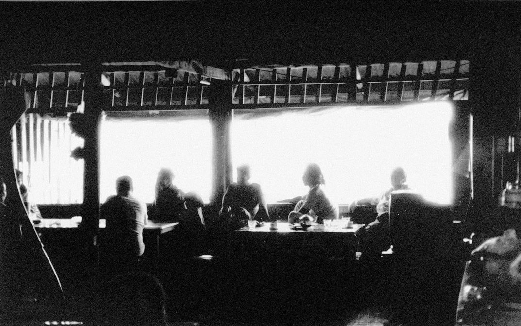 Film still from MYSTERIOUS OBJECT AT NOON: A slightly blurred shot of a group of people sitting at a long table. Bright light streams into the room through a window.