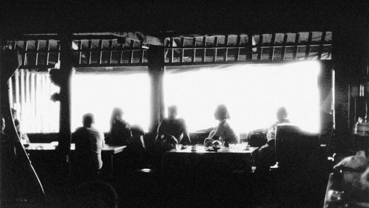  Film still from MYSTERIOUS OBJECT AT NOON: A slightly blurred shot of a group of people sitting at a long table. Bright light streams into the room through a window.