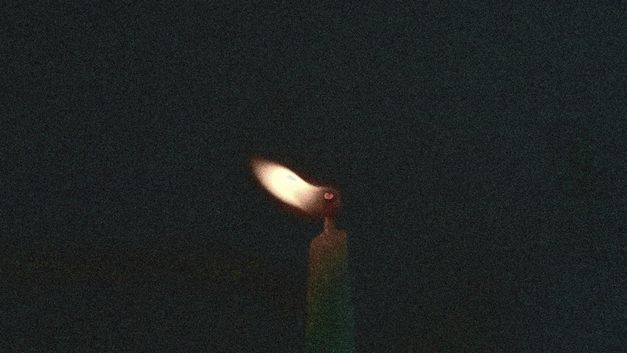 Film still from Mary Helena Clark’s „Exhibition“. A grainy picture of the flame of a candle in the dark.
