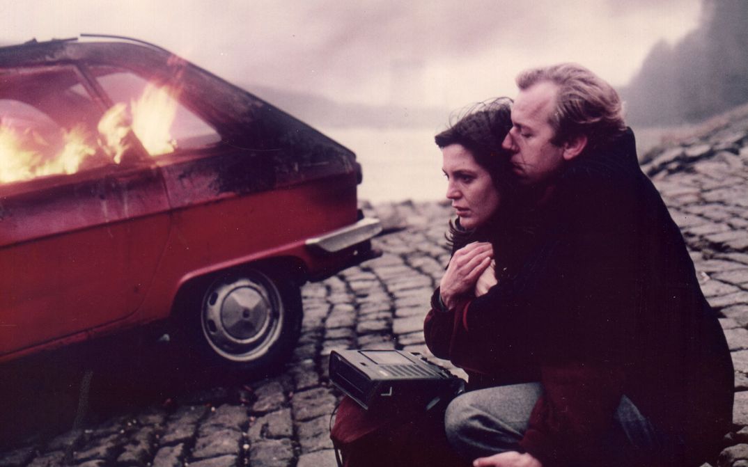 Filmstill aus LETZTE LIEBE. Ein Paar mit einem Kassettenrekorder auf der Straße, im Hintergrund ein brennendes Auto..
