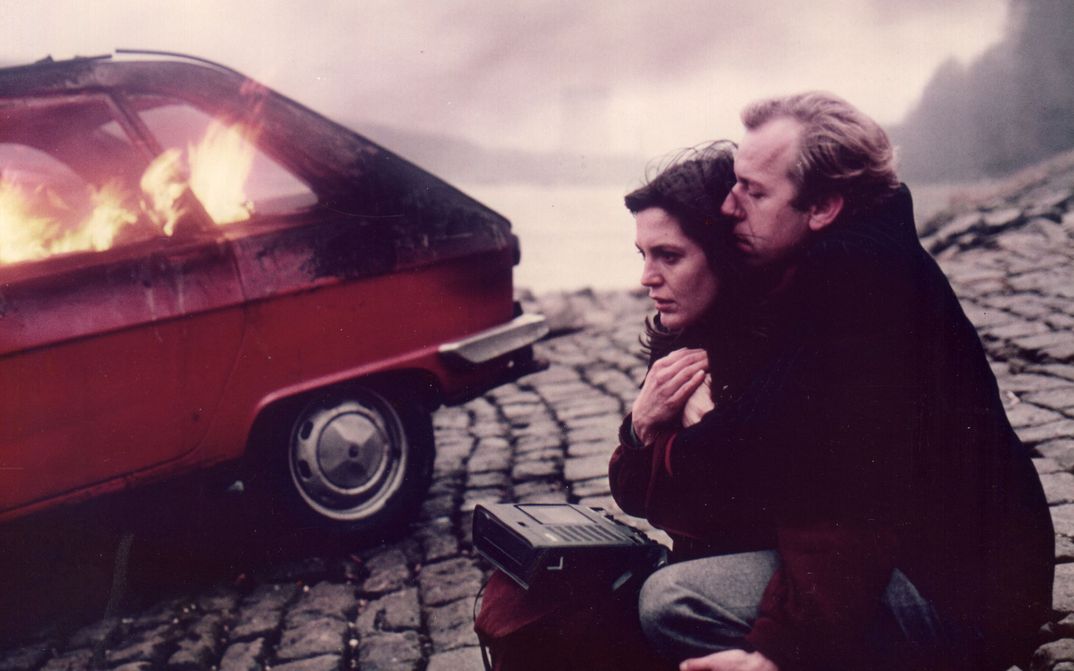 Filmstill aus LETZTE LIEBE. Ein Paar mit einem Kassettenrekorder auf der Straße, im Hintergrund ein brennendes Auto..