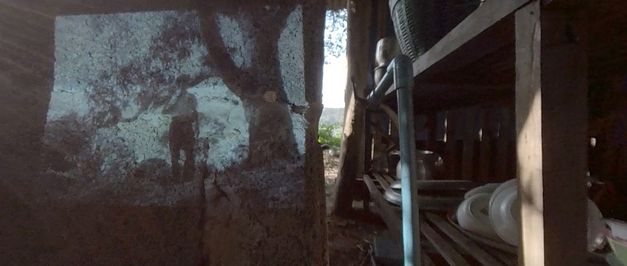 Filmstill aus dem Film "Trip After" von Ukrit Sa-nguanhai. Der Ausschnitt einer Kochstelle, ein Wasserhahn und - auf eine Holzwand projiziert - das Bild einer älteren Person unter einem Baum.