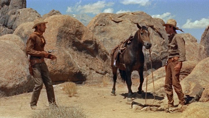 Film still from RIDE LONESOME: Two cowboys face each other in a rocky desert landscape, with a horse standing between them. One is pointing his revolver at the other.