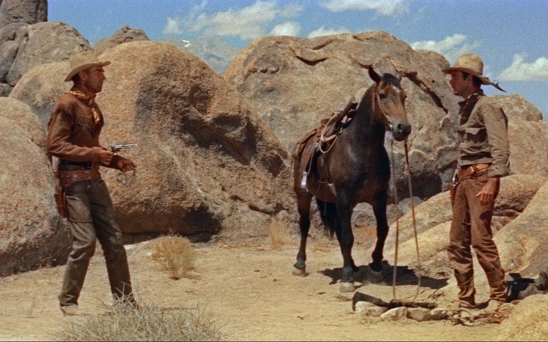 Filmstill aus RIDE LONESOME: Zwei Cowboys stehen sich in einer felsigen Wüstenlandschaft gegenüber, zwischen ihnen steht ein Pferd. Einer richtet seinen Revolver auf den anderen.