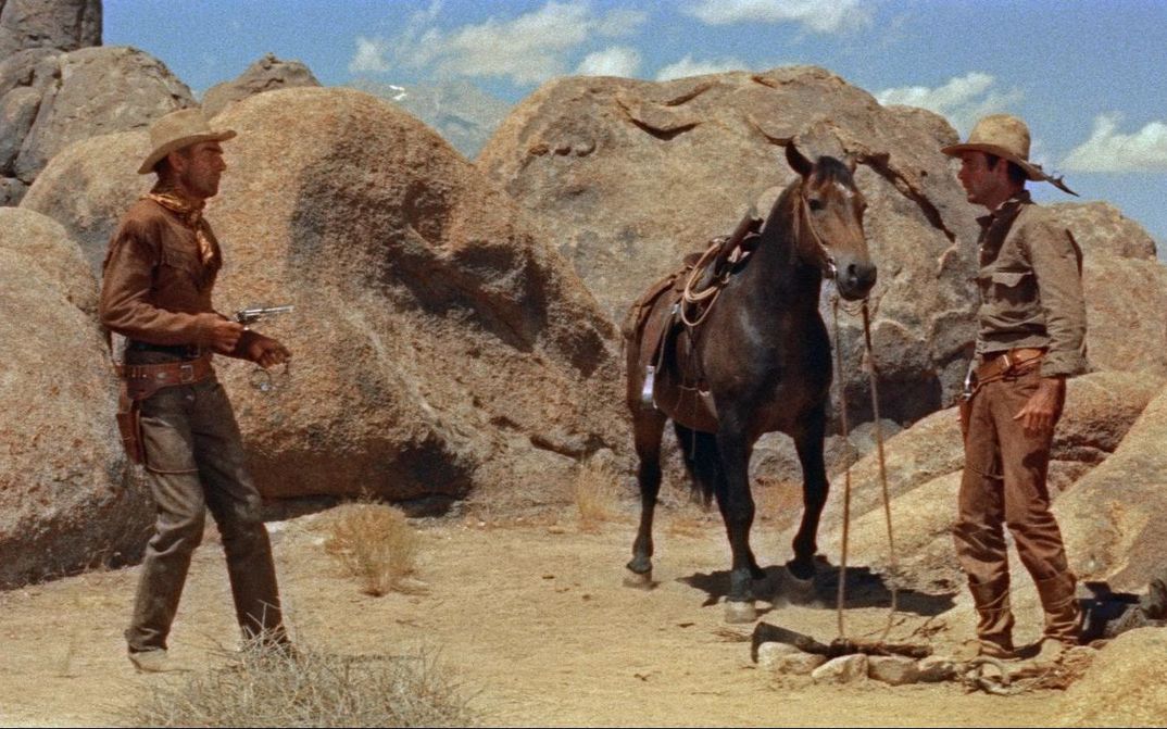 Filmstill aus RIDE LONESOME: Zwei Cowboys stehen sich in einer felsigen Wüstenlandschaft gegenüber, zwischen ihnen steht ein Pferd. Einer richtet seinen Revolver auf den anderen.