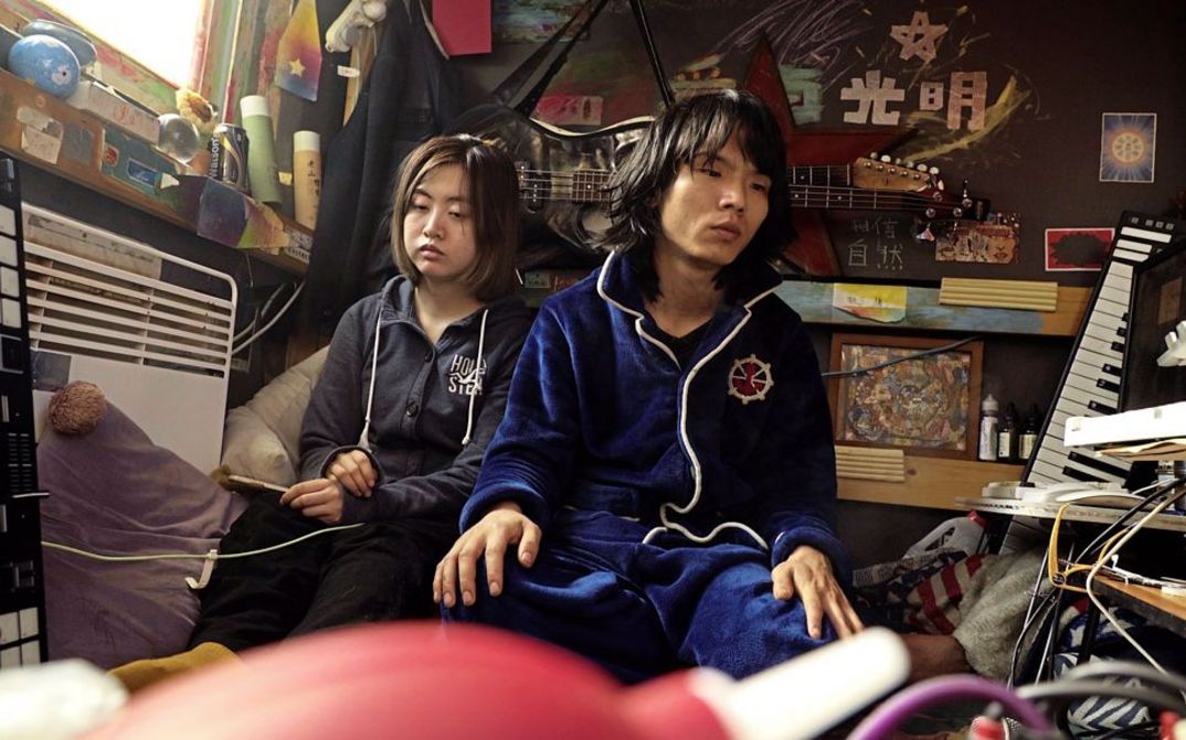 Film still from "Republic" by Jin Jiang. It shows two people sitting against the wall in a crowded little room. 