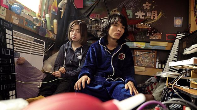 Film still from "Republic" by Jin Jiang. It shows two people sitting against the wall in a crowded little room. 