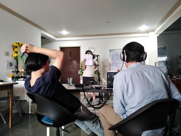 A behind-the-scenes shot of three people. Two are sitting at a desk as the other one stands across from them holding a white sheet of paper in front of their face.