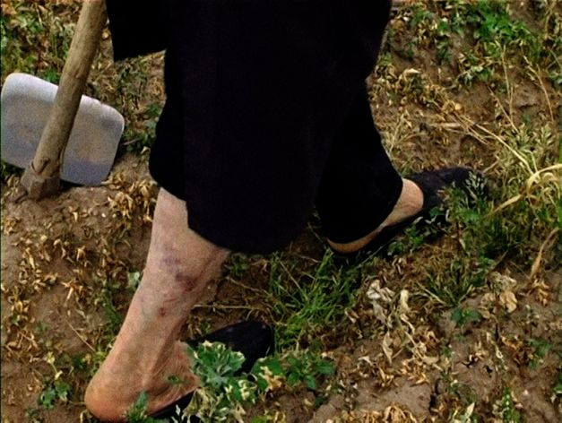 Still from the film "Terra que marca (Striking Land)" by Raul Domingues. Close-up of the legs of a women farmer walking through the field. In the left hand corner we see a hoe. 