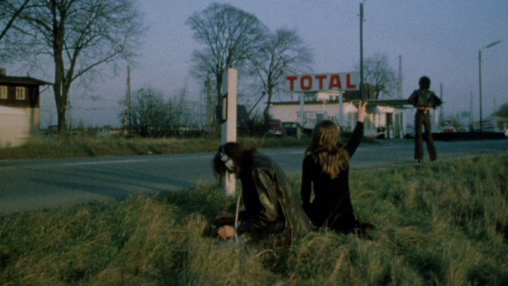Film still from LEAVE ME ALONE. Three people are standing on the side of a road, with a gas station in the background.