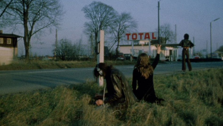 Filmstill aus LEAVE ME ALONE. Drei Menschen stehen am Rand einer Straße, im Hintergrund eine Tankstelle.