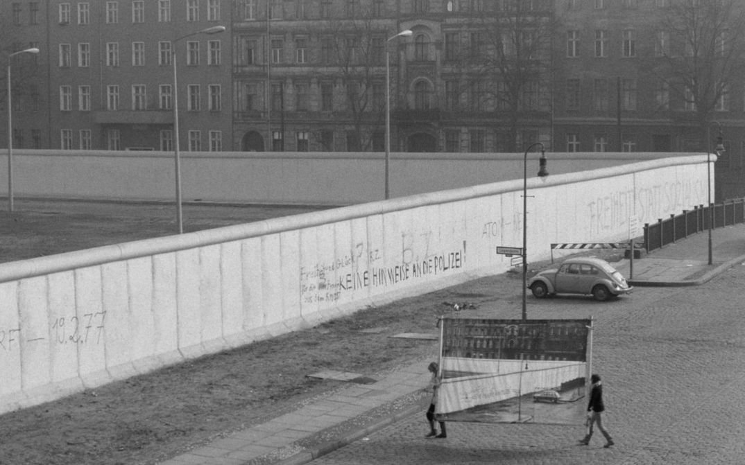 Film still from REDUPERS: View of the Berlin Wall in Kreuzberg. Small, you can see two women carrying a photo wall with this very view.