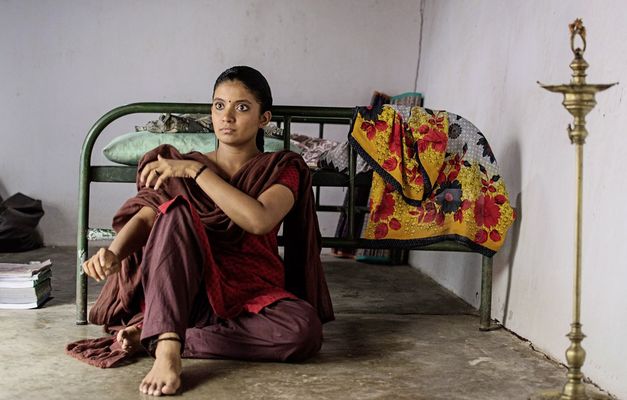 Film still from "Kottukkaali" by Vinothraj PS. It shows a woman with braided hair and a dot between her eyebrows sitting on the floor with her back to a bed. 