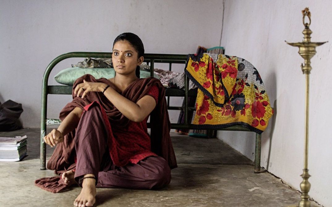 Filmstill aus "Kottukkaali" von Vinothraj PS. Zu sehen ist eine Frau mit geflochtenem Haar und einem Punkt zwischen den Augenbrauen, die mit dem Rücken zu einem Bett auf dem Boden sitzt. 