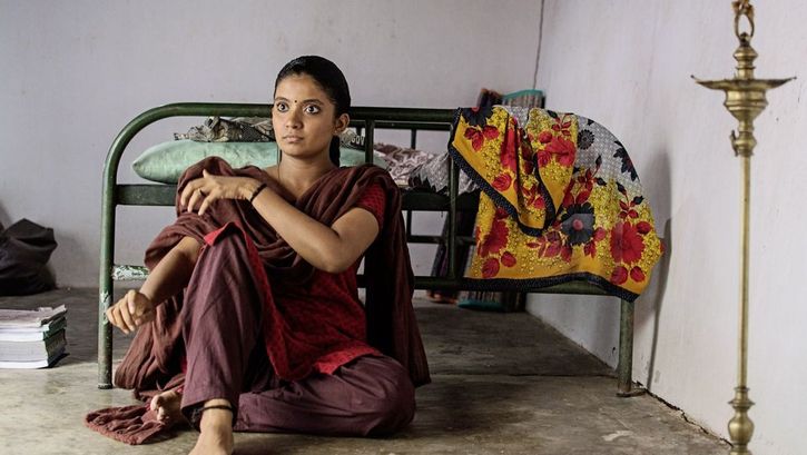 Film still from "Kottukkaali" by Vinothraj PS. It shows a woman with braided hair and a dot between her eyebrows sitting on the floor with her back to a bed. 