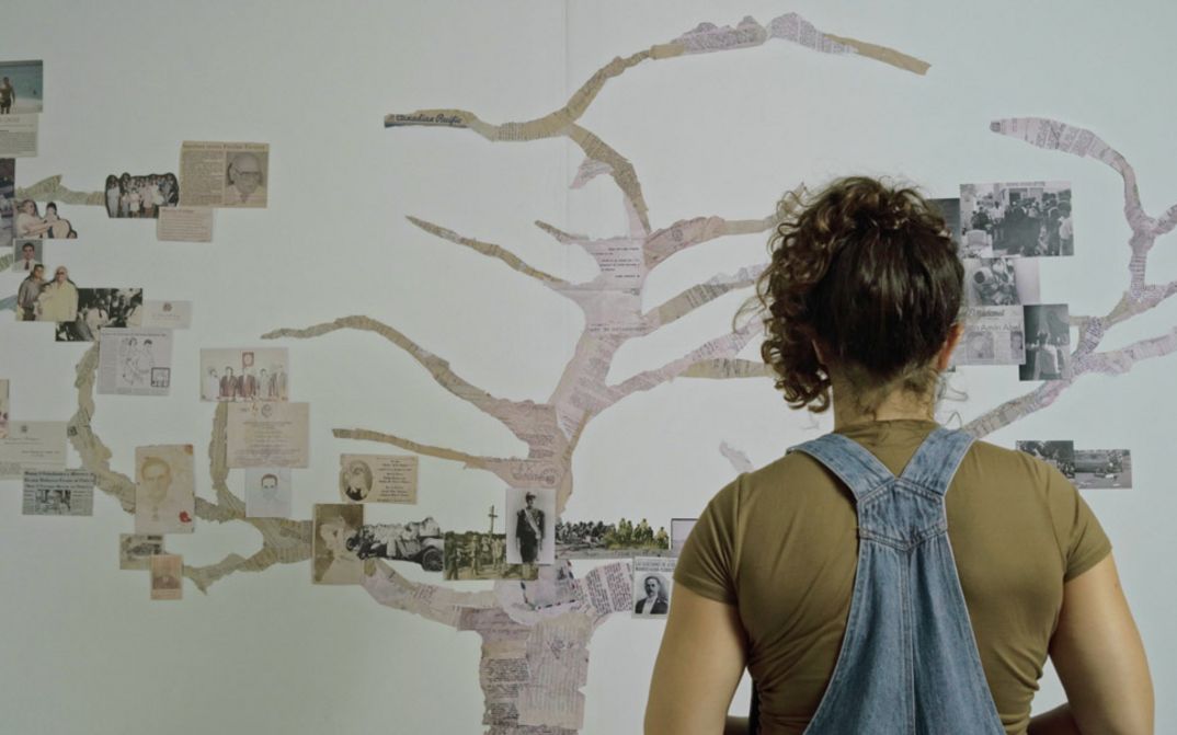 Film still from COLOSAL by Nayibe Tavares-Abel. The image shows a woman looking at a tree made of newspaper clippings and photos glued to the wall.