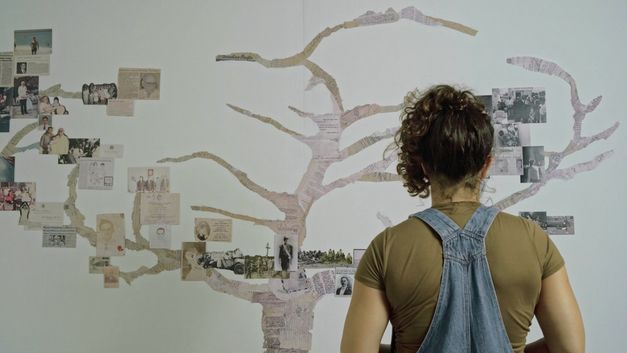 Filmstill aus COLOSAL von Nayibe Tavares-Abel. Das Bild zeigt eine Frau die auf einen Baum schaut, der aus Zeitungauschnitten und Fotos auf die Wand geklebt ist. 