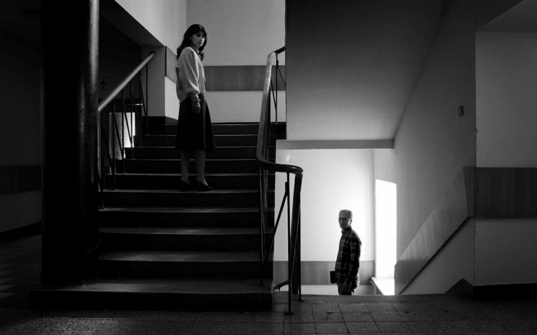 Film still from Assaf Gruber’s film “Miraculous Accident”. On a staircase filmed frontally in black and white, two people face the camera: a woman on the ascending flight, and an older man on the descending one.