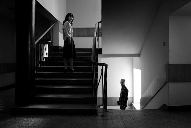 Film still from Assaf Gruber’s film “Miraculous Accident”. On a staircase filmed frontally in black and white, two people face the camera: a woman on the ascending flight, and an older man on the descending one.
