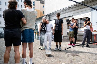 A behind-the-scenes shot with eight people. One of them is filming, and the others stand around a metal pot with steam coming out of it.